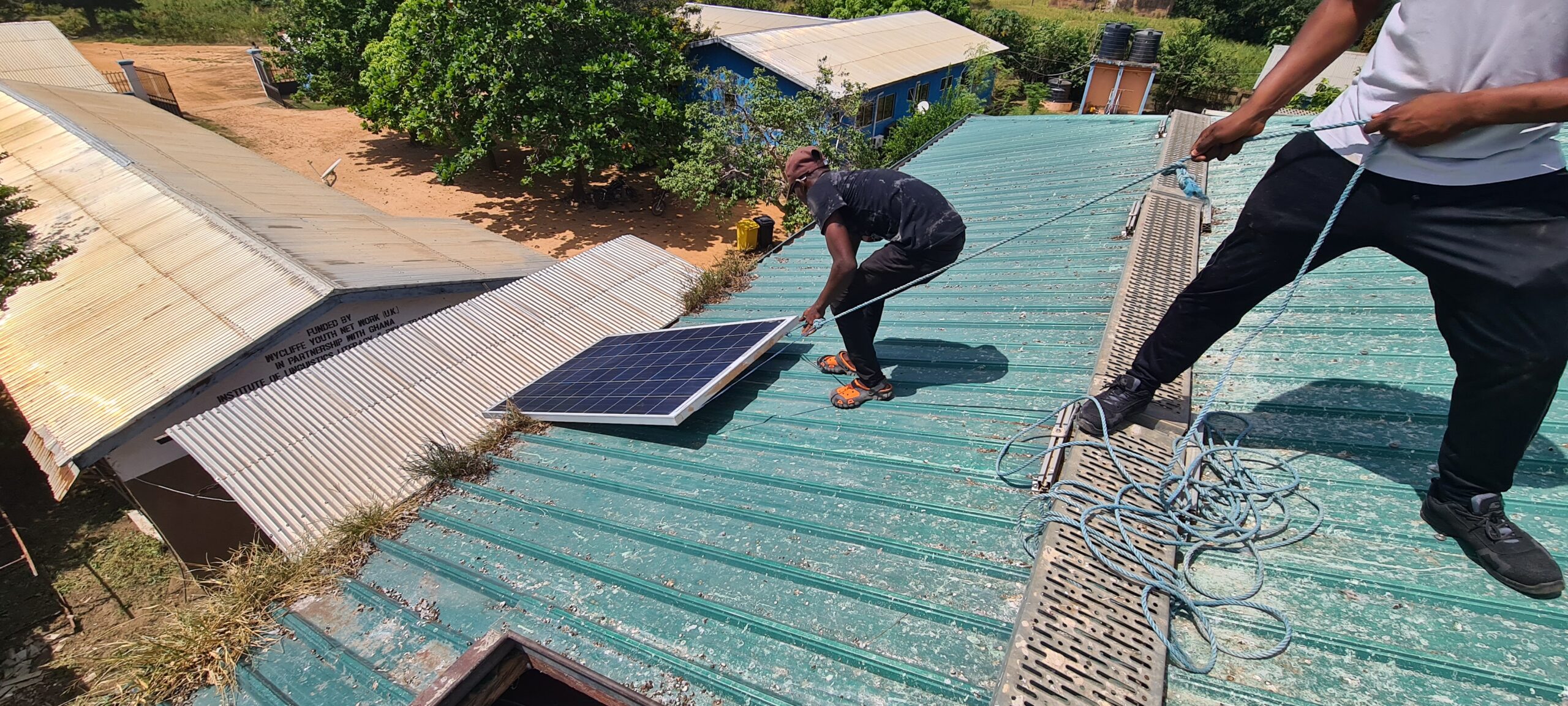 Zwei lokale Installateure ziehen ein Solarpanel auf das Dach des SILAP Guesthouse, während das Dach mit Taubenkot übersät ist.
