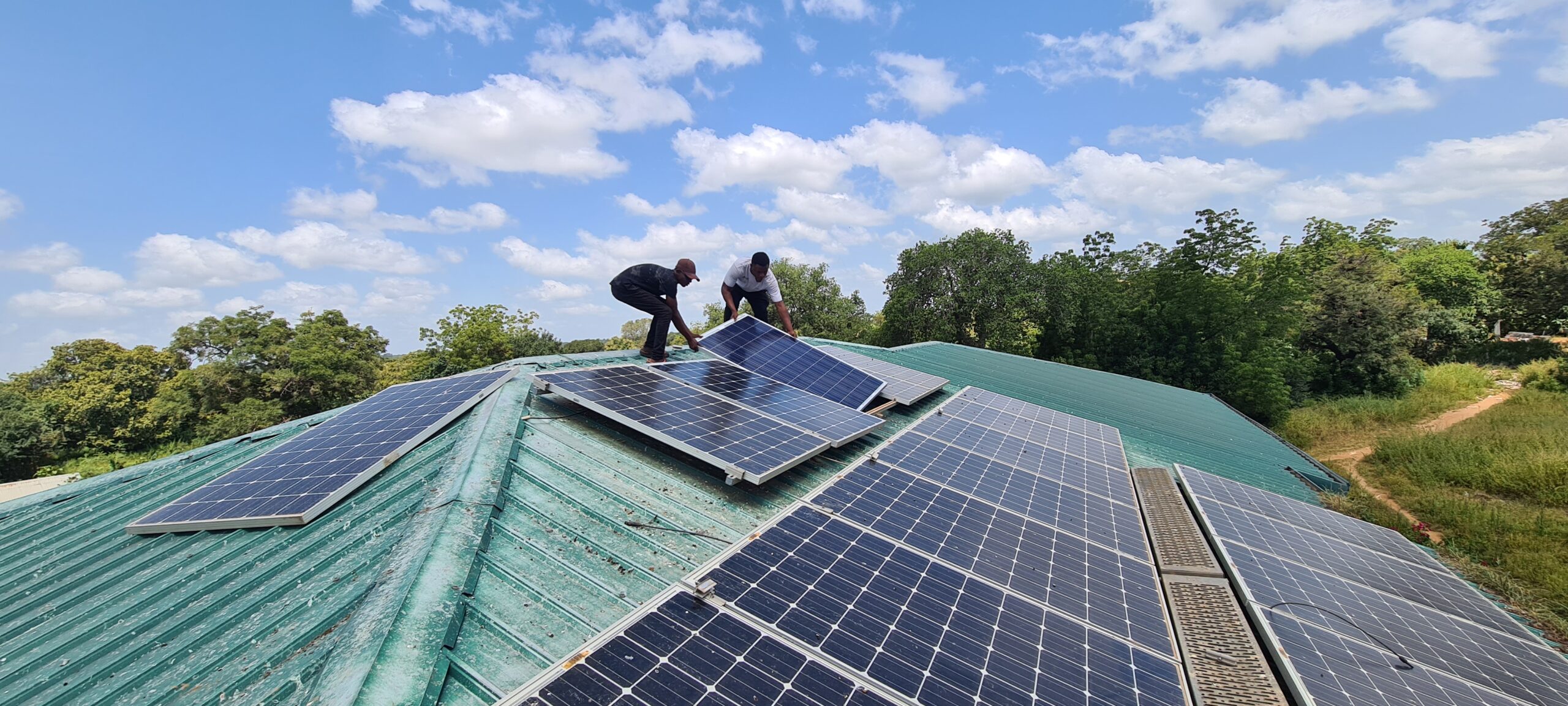 Zwei lokale Installateure installieren ein neues Solarpanel auf dem Dach des Gästehauses, während ein weiteres ungesichertes Panel daneben liegt.