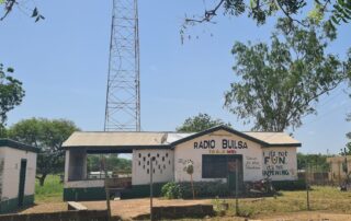 Hausfront von Radio Builsa mit dem Schriftzug „Radio Builsa“ in Schwarz und „106.5 MHz“ in Gelb und Rot über der Tür. Rechts steht der Spruch „If it's not FUN it's not HAPPENING“ in fröhlichen Farben (Schwarz, Gelb, Grün). Im Hintergrund ist der Sendemast teilweise sichtbar. Das Bild erscheint im News-Beitrag Radio Builsa's Solar Repairs