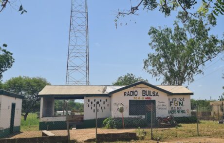 Hausfront von Radio Builsa mit dem Schriftzug „Radio Builsa“ in Schwarz und „106.5 MHz“ in Gelb und Rot über der Tür. Rechts steht der Spruch „If it's not FUN it's not HAPPENING“ in fröhlichen Farben (Schwarz, Gelb, Grün). Im Hintergrund ist der Sendemast teilweise sichtbar. Das Bild erscheint im News-Beitrag Radio Builsa's Solar Repairs