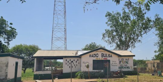 Hausfront von Radio Builsa mit dem Schriftzug „Radio Builsa“ in Schwarz und „106.5 MHz“ in Gelb und Rot über der Tür. Rechts steht der Spruch „If it's not FUN it's not HAPPENING“ in fröhlichen Farben (Schwarz, Gelb, Grün). Im Hintergrund ist der Sendemast teilweise sichtbar. Das Bild erscheint im News-Beitrag Radio Builsa's Solar Repairs