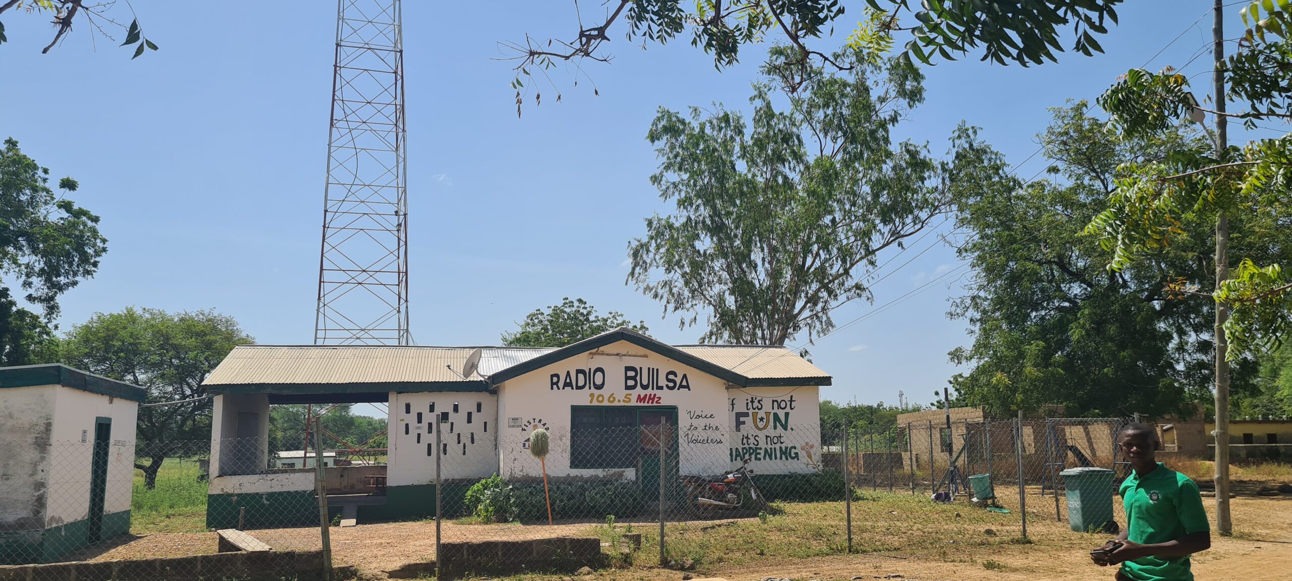 Hausfront von Radio Builsa mit dem Schriftzug „Radio Builsa“ in Schwarz und „106.5 MHz“ in Gelb und Rot über der Tür. Rechts steht der Spruch „If it's not FUN it's not HAPPENING“ in fröhlichen Farben (Schwarz, Gelb, Grün). Im Hintergrund ist der Sendemast teilweise sichtbar. Das Bild erscheint im News-Beitrag Radio Builsa's Solar Repairs