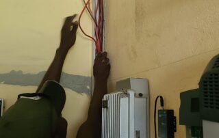 Ein Installateur steht vor den neu installierten Geräten der PV-Anlage von Radio Builsa, mit den Händen in den Kabeln des offenen Kabelkanals.