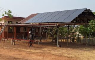 Ein offener Hausteil der GfC-Bimbilla Mission mit einer PV-Anlage auf dem Dach. Im Vordergrund bearbeitet ein Arbeiter ein Metallstück, während der trockene, braune Boden die Umgebung prägt.