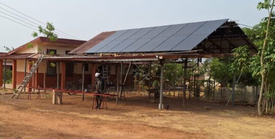 Ein offener Hausteil der GfC-Bimbilla Mission mit einer PV-Anlage auf dem Dach. Im Vordergrund bearbeitet ein Arbeiter ein Metallstück, während der trockene, braune Boden die Umgebung prägt.