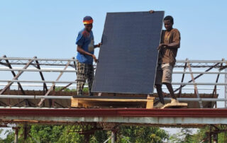 Zwei Ghanaer legen das erste PV-Panel in die Alu-Schienen auf dem Dach. Sie stehen auf Holzbrettern, die behelfsmäßig als Gerüst dienen.