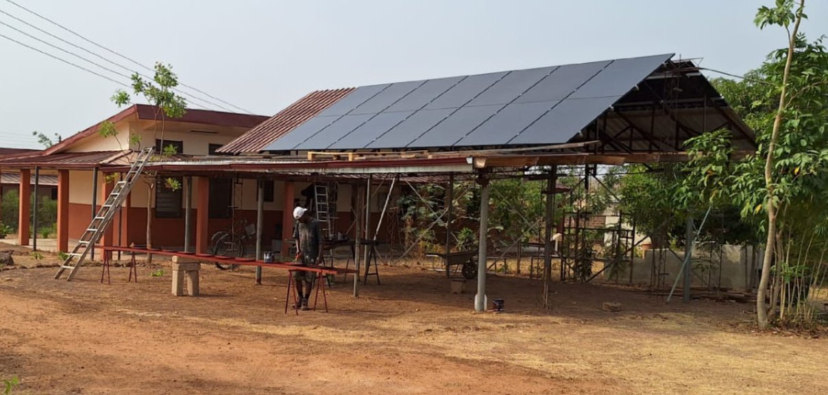 Ein offener Hausteil der GfC-Bimbilla Mission mit einer PV-Anlage auf dem Dach. Im Vordergrund bearbeitet ein Arbeiter ein Metallstück, während der trockene, braune Boden die Umgebung prägt.