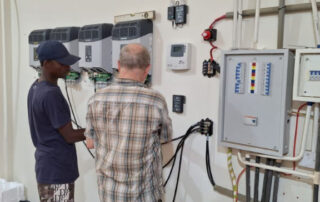 Die drei Wechselrichter und der Ladeinverter werden angeschlossen, daneben die Kommunikationsmodule und ein Schaltschrank mit verschiedenen dicken Stromleitungen. Werner und Attah arbeiten daran, alles zu verkabeln.