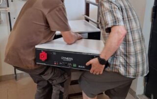 Werner und Roland heben gemeinsam eine der neuen Batterien ins Batteriegestell. Man sieht, wie schwer die Batterie ist, und sie schwitzen vor Anstrengung.